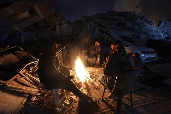مردم چهارشنبه 8 فوریه 2023 در مقابل ساختمان های ویران شده در آنتاکیا، جنوب ترکیه،  خود را با آتش گرم می کنند.  - اسپوتنیک ایران  