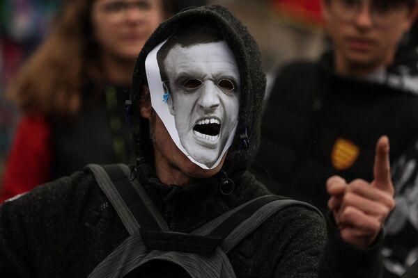 یک معترض که ماسکی شبیه به رئیس‌جمهور فرانسه بر تن دارد، هنگام شرکت در تظاهرات روز اول ماه مه (روز کارگر) به مناسبت روز جهانی کارگران. بوردو، جنوب غربی فرانسه، اول مه 2023. - اسپوتنیک ایران  