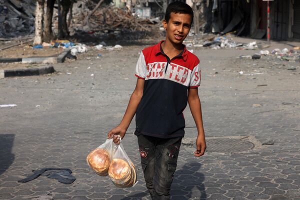 پسر جوانی کیسه‌های نان را حمل می‌کند در حالی که از مقابل ساختمان آسیب دیده در اثر حملات اسرائیل در شهر غزه عبور می کند. - اسپوتنیک ایران  