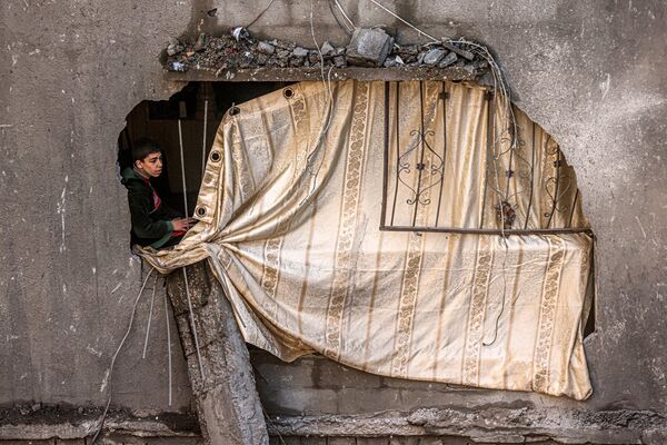 پسری در کنار سوراخ دیوار پوشیده شده با پرده در ساختمانی مشرف به ساختمانی که در اثر بمباران اسرائیل در رفح در جنوب نوار غزه ویران شده، نشسته است. - اسپوتنیک ایران  