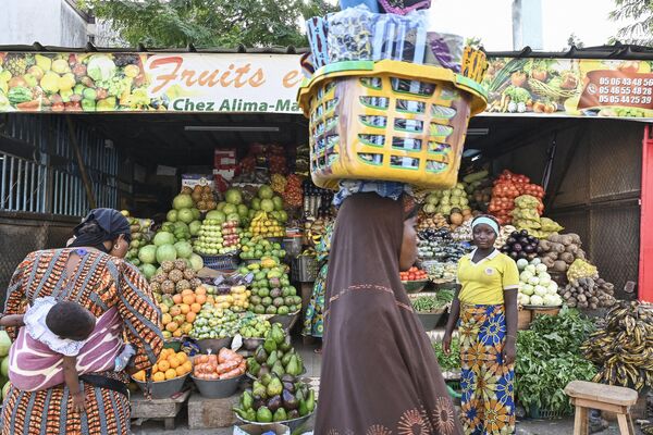 زنی سبد بر سر در حال عبور از کنار زنی میوه و سبزی فروش در ماه مبارک رمضان در ابیجان. - اسپوتنیک ایران  