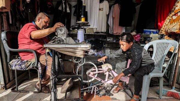 دختربچه ای در شهر رفح،  دوچرخه‌ ای را پدال می‌زند که به منبع موقتی برق برای خیاطی که با چرخ خیاطی کار می‌کند، تبدیل شده است - اسپوتنیک ایران  