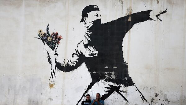 Two men are sitting in front of a famous graffiti of British street artist Banksy, painted on a wall of a gas station in the West Bank city of Bethlehem on December 16, 2015. - اسپوتنیک ایران  