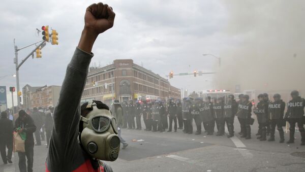 A demonstrator raises his fist as police stand in formation as a store burns, Monday, April 27, 2015, during unrest following the funeral of Freddie Gray in Baltimore - اسپوتنیک ایران  
