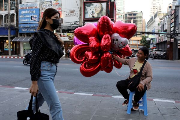 زنی با ماسک از کنار فروشنده بادکنک ولنتاین در یکی از خیابان های بانکوک تایلند عبور می کند - اسپوتنیک ایران  